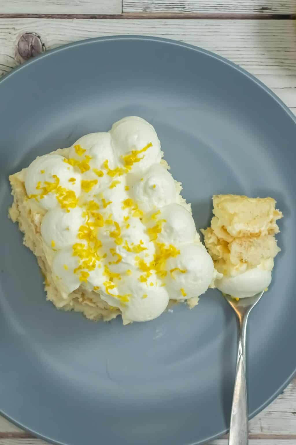 A slice of Limoncello Tiramisu topped with whipped cream and lemon zest sits on a gray plate, with a fork holding a bite-sized portion.