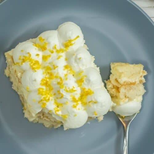 A slice of Limoncello Tiramisu topped with whipped cream and lemon zest sits on a gray plate, with a fork holding a bite-sized portion.