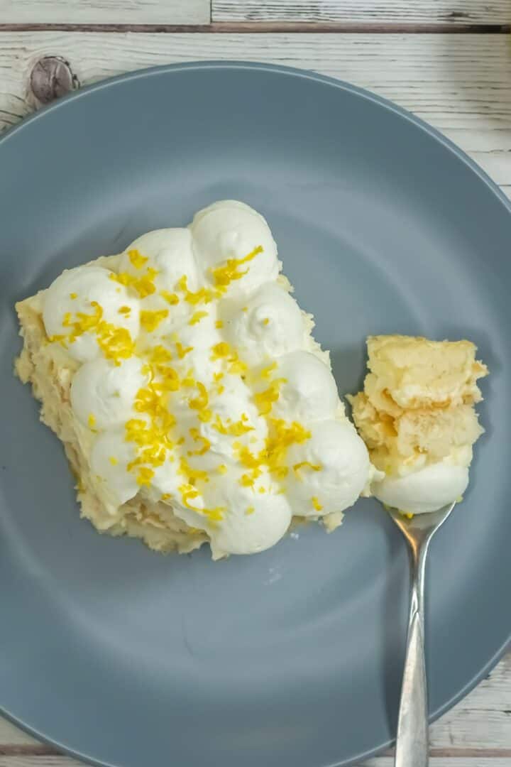 A slice of Limoncello Tiramisu topped with whipped cream and lemon zest sits on a gray plate, with a fork holding a bite-sized portion.