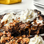 A decadent chocolate brownie pie topped with caramel, pecans, and whipped cream on a glass plate.