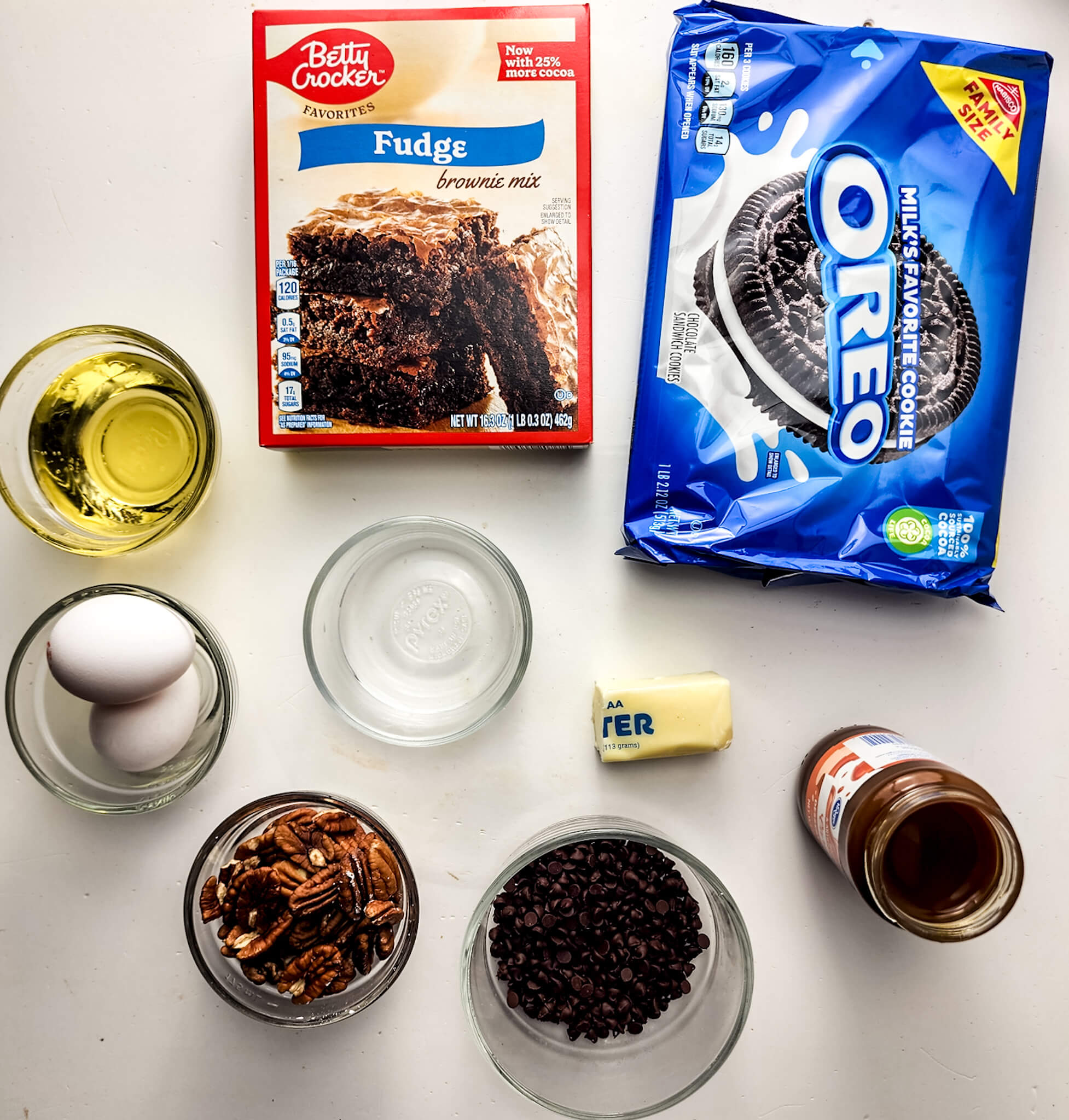 Ingredients for baking a brownie pie laid out on a white surface, including a box of fudge brownie mix, oreo cookies, oil, eggs, pecans, butter, chocolate chips,