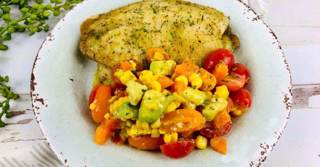 A seasoned baked haddock fillet served with a colorful vegetable medley of corn, avocado, tomatoes, and carrots on a white rustic plate.