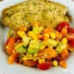 A seasoned baked haddock fillet served with a colorful vegetable medley of corn, avocado, tomatoes, and carrots on a white rustic plate.