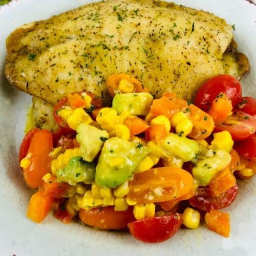 A seasoned baked haddock fillet served with a colorful vegetable medley of corn, avocado, tomatoes, and carrots on a white rustic plate.