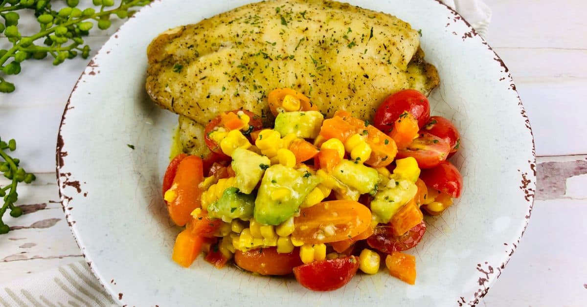 A seasoned baked haddock fillet served with a colorful vegetable medley of corn, avocado, tomatoes, and carrots on a white rustic plate.