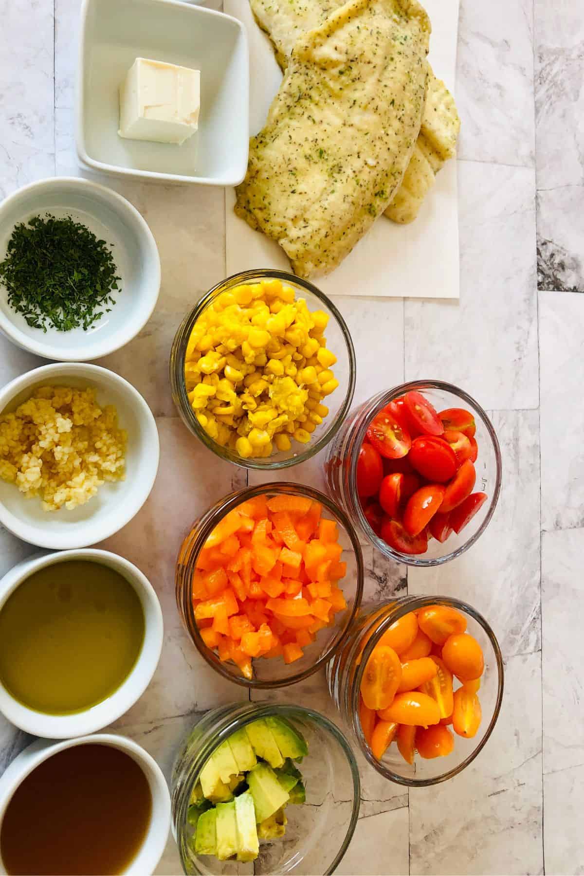 Various ingredients in bowls, including baked  fillets, cherry tomatoes, orange bell pepper, corn, avocado, herbs, minced garlic, butter, olive oil, and vinegar, arranged on a countertop.