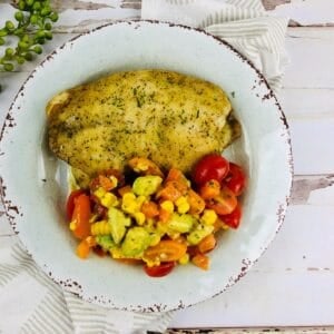 A plate with a seasoned baked haddock fillet and a side of chopped tomato, corn, avocado, and bell pepper salad.