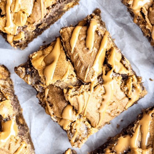 Close-up of biscoff brownies drizzled with caramel, scattered with chocolate chips, arranged in a grid pattern on parchment paper.