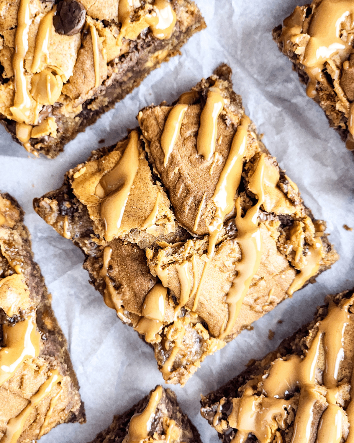 Close-up of biscoff brownies drizzled with caramel, scattered with chocolate chips, arranged in a grid pattern on parchment paper.