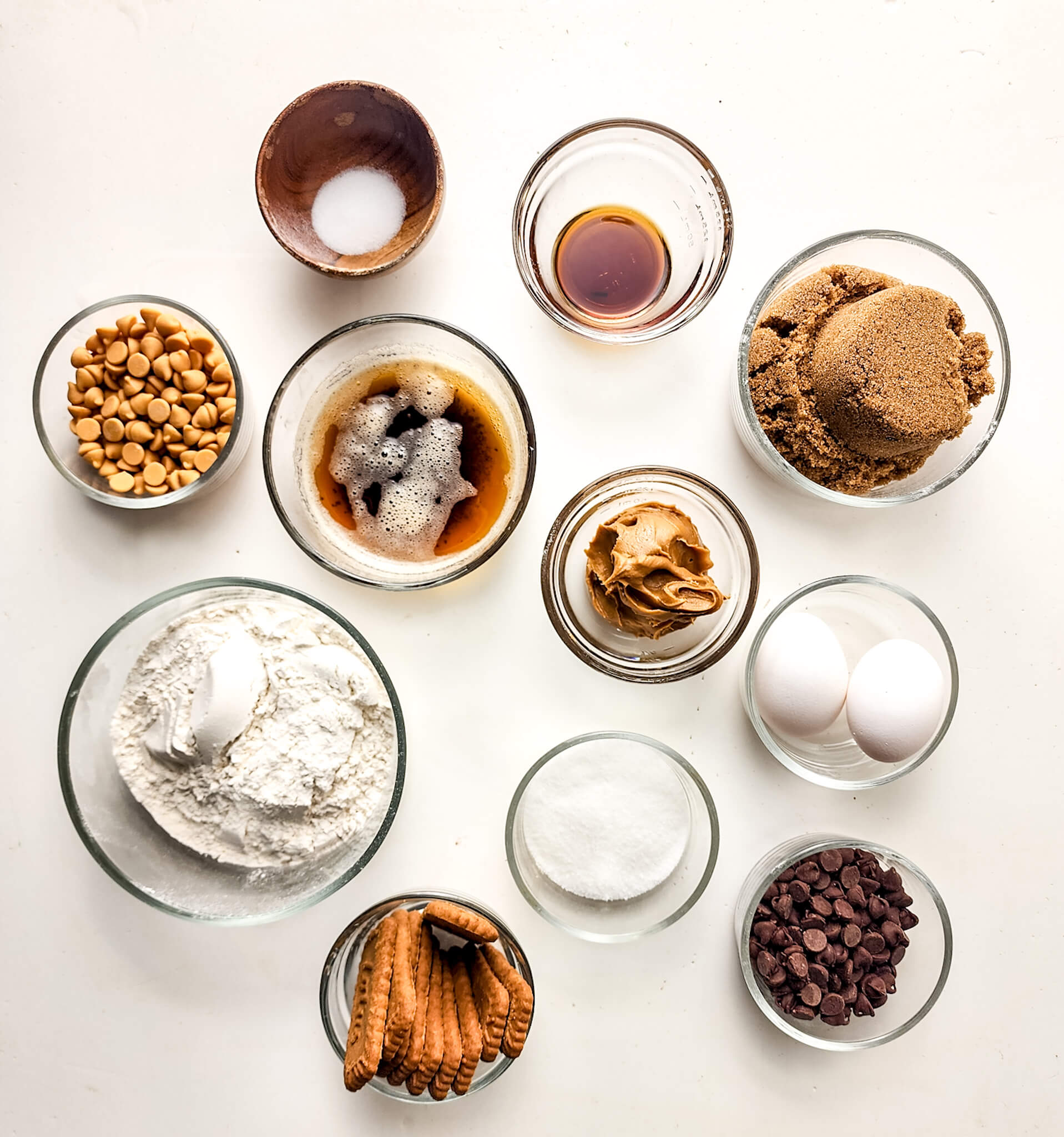 Various baking ingredients neatly arranged in clear bowls on a white surface, including flour, eggs, sugar, nuts, and Biscoff brownies.