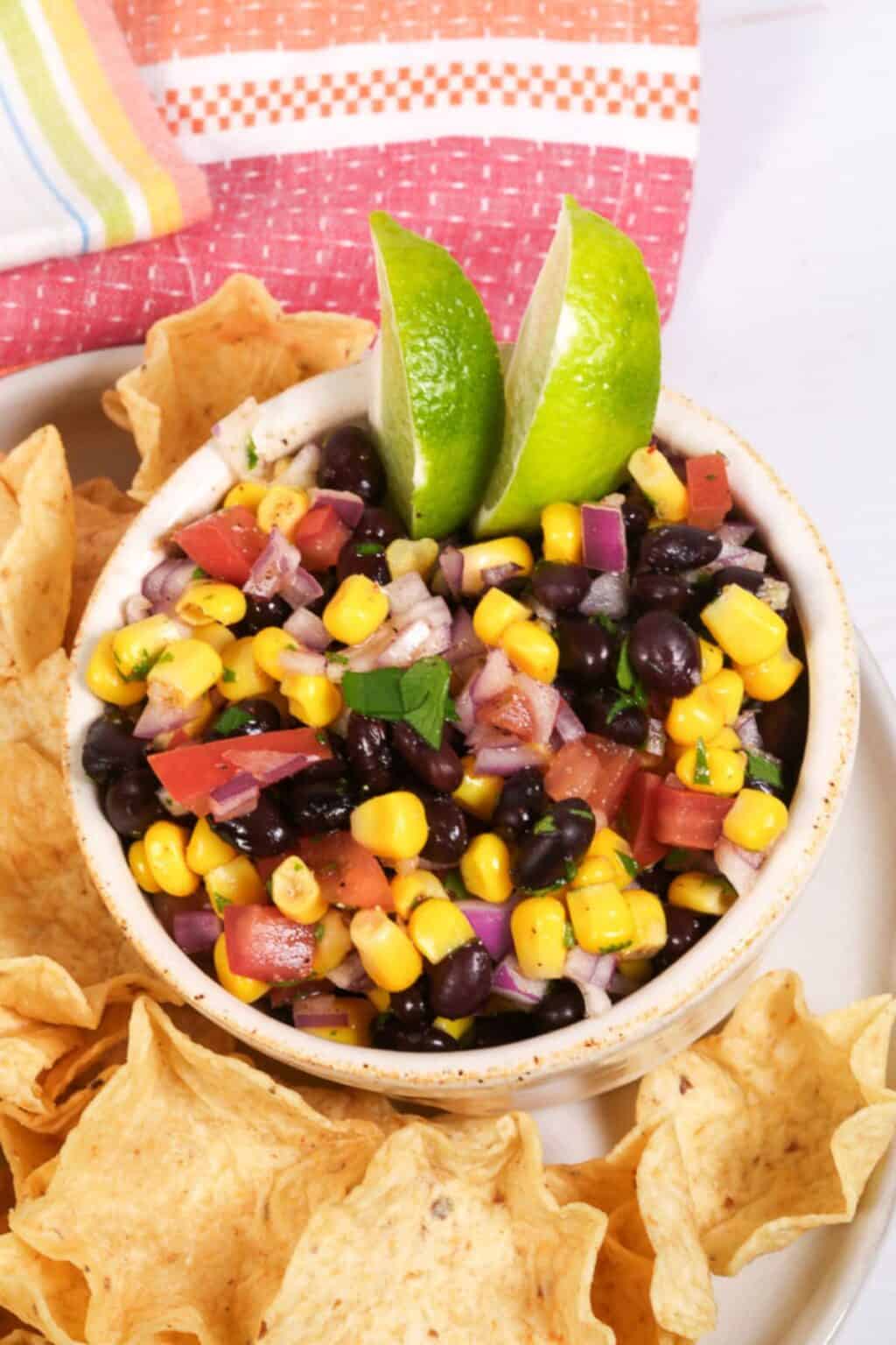 A bowl of Black Bean and Corn Salsa with chopped tomatoes, red onion, and herbs, garnished with lime wedges and surrounded by crispy tortilla chips.
