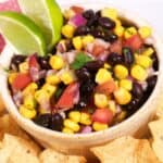 A bowl of Black Bean and Corn Salsa with diced tomatoes, onions, and cilantro, garnished with lime wedges and surrounded by crispy tortilla chips.