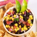 A bowl of Black Bean and Corn Salsa with diced tomatoes and onions, garnished with lime wedges, surrounded by crisp tortilla chips.