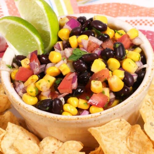 A bowl of Black Bean and Corn Salsa with diced tomatoes, onions, and cilantro, garnished with lime wedges and surrounded by crispy tortilla chips.