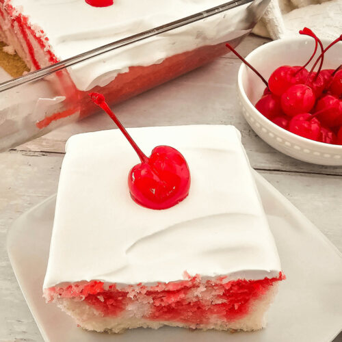 A slice of Cherry Poke Cake with a creamy white topping and a single cherry garnish, served on a white plate.