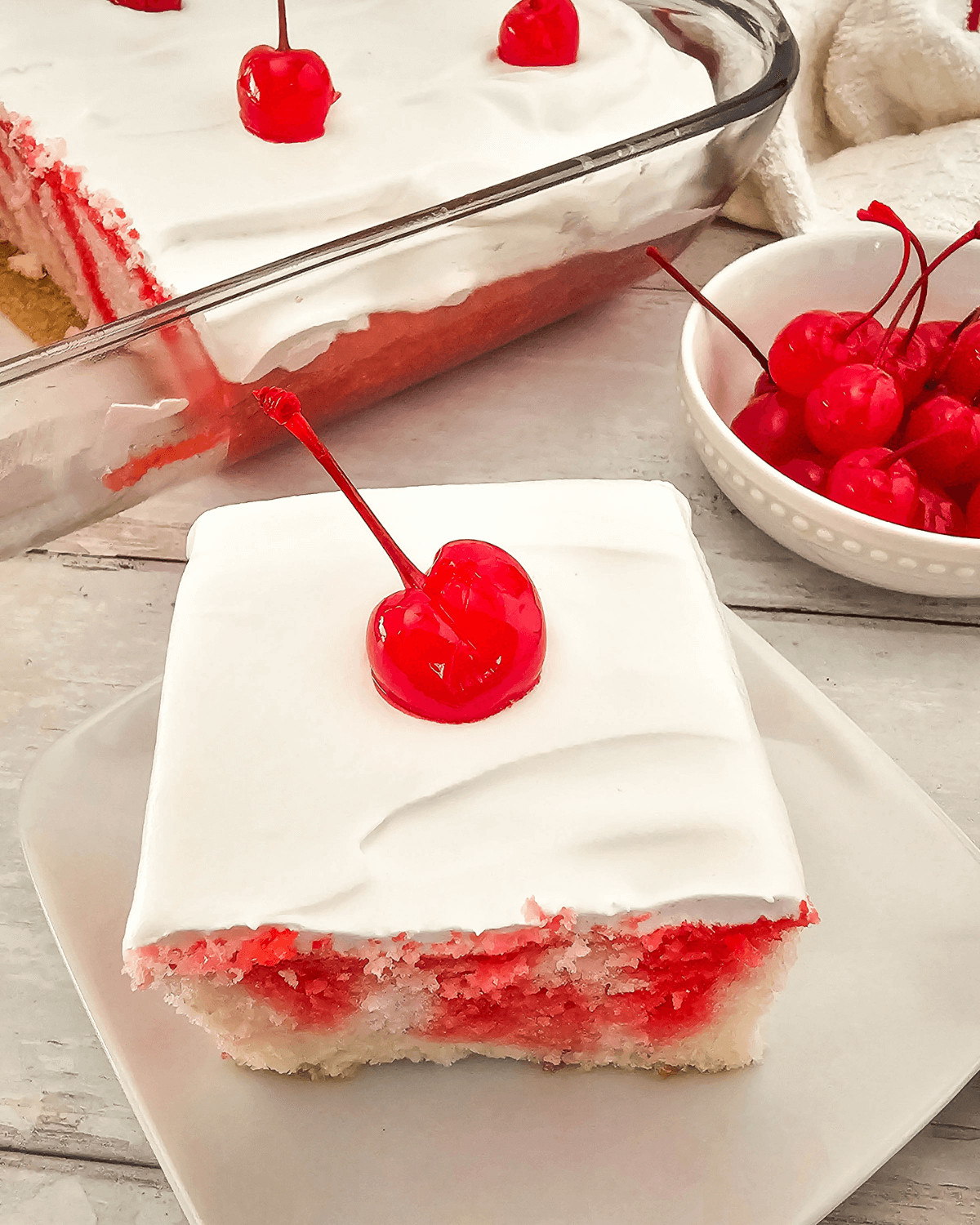 A slice of Cherry Poke Cake with a creamy white topping and a single cherry garnish, served on a white plate.