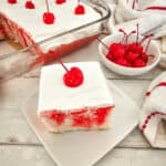 A slice of cherry poke cake on a white plate, with a glass dish of cherry poke cake and a bowl of cherries in the background.