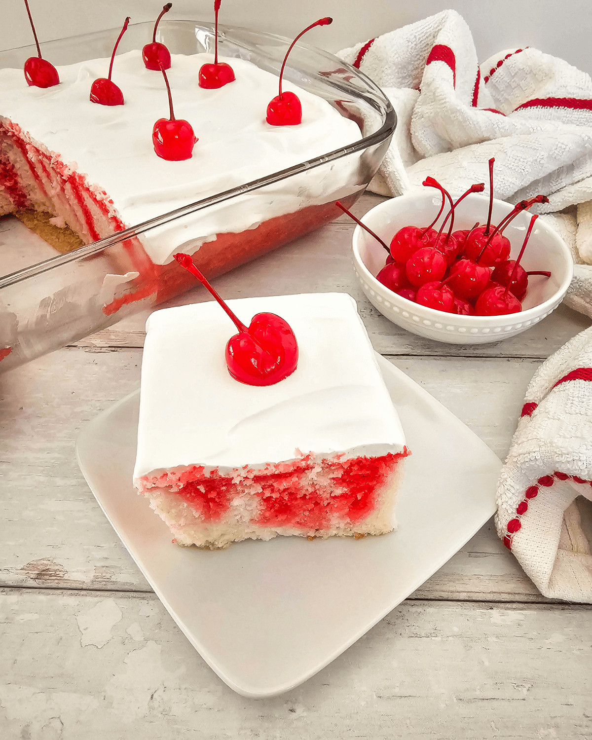 A slice of cherry poke cake on a white plate, with a glass dish of cherry poke cake and a bowl of cherries in the background.