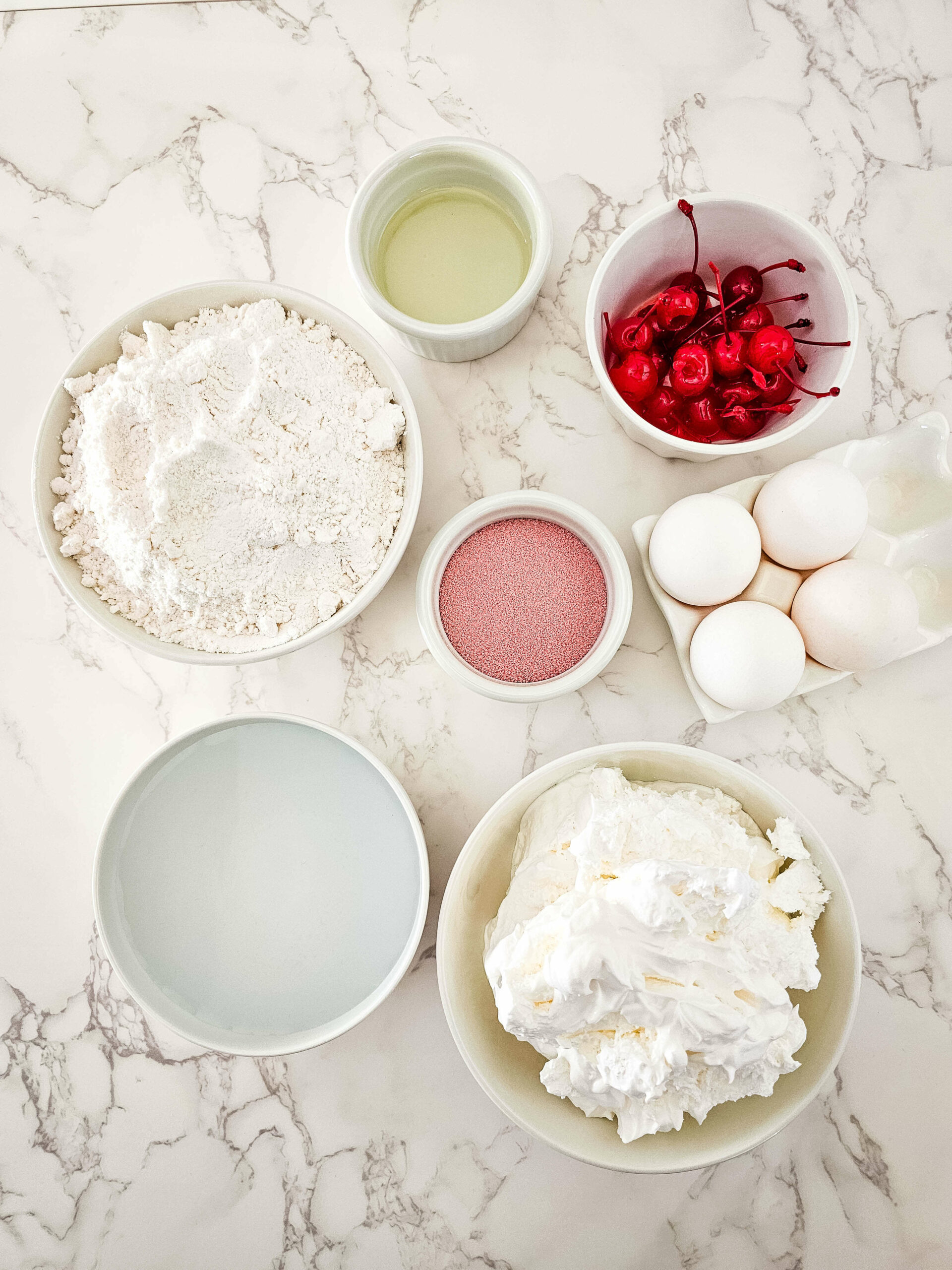 Various ingredients for the dessert arranged on a marble surface, including flour, eggs, cherries, oil, and whipped cream.