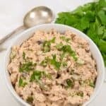 A white bowl filled with chipotle chicken salad garnished with chopped herbs, placed next to a bunch of fresh cilantro and a spoon on a white surface.