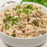 A bowl of chipotle chicken salad garnished with chopped parsley, placed on a white surface with fresh herbs and a spoon nearby.