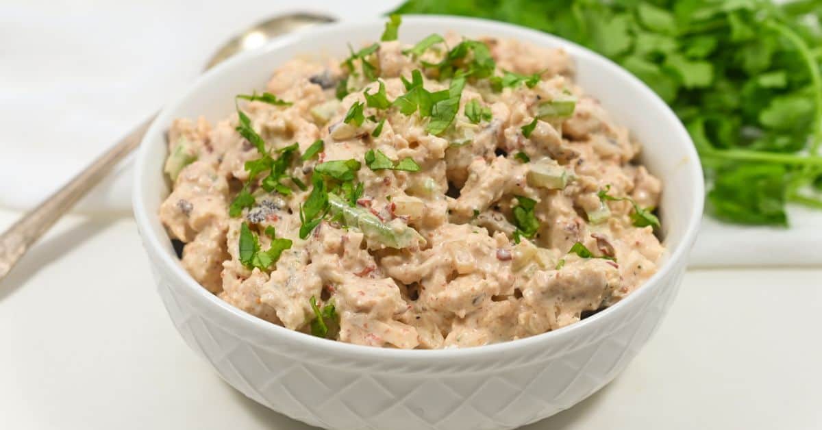 A bowl of chipotle chicken salad garnished with chopped parsley, placed on a white surface with fresh herbs and a spoon nearby.