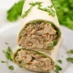 Two halves of a chipotle chicken salad wrap stacked on a white plate, garnished with chopped parsley, with leafy greens in the background.