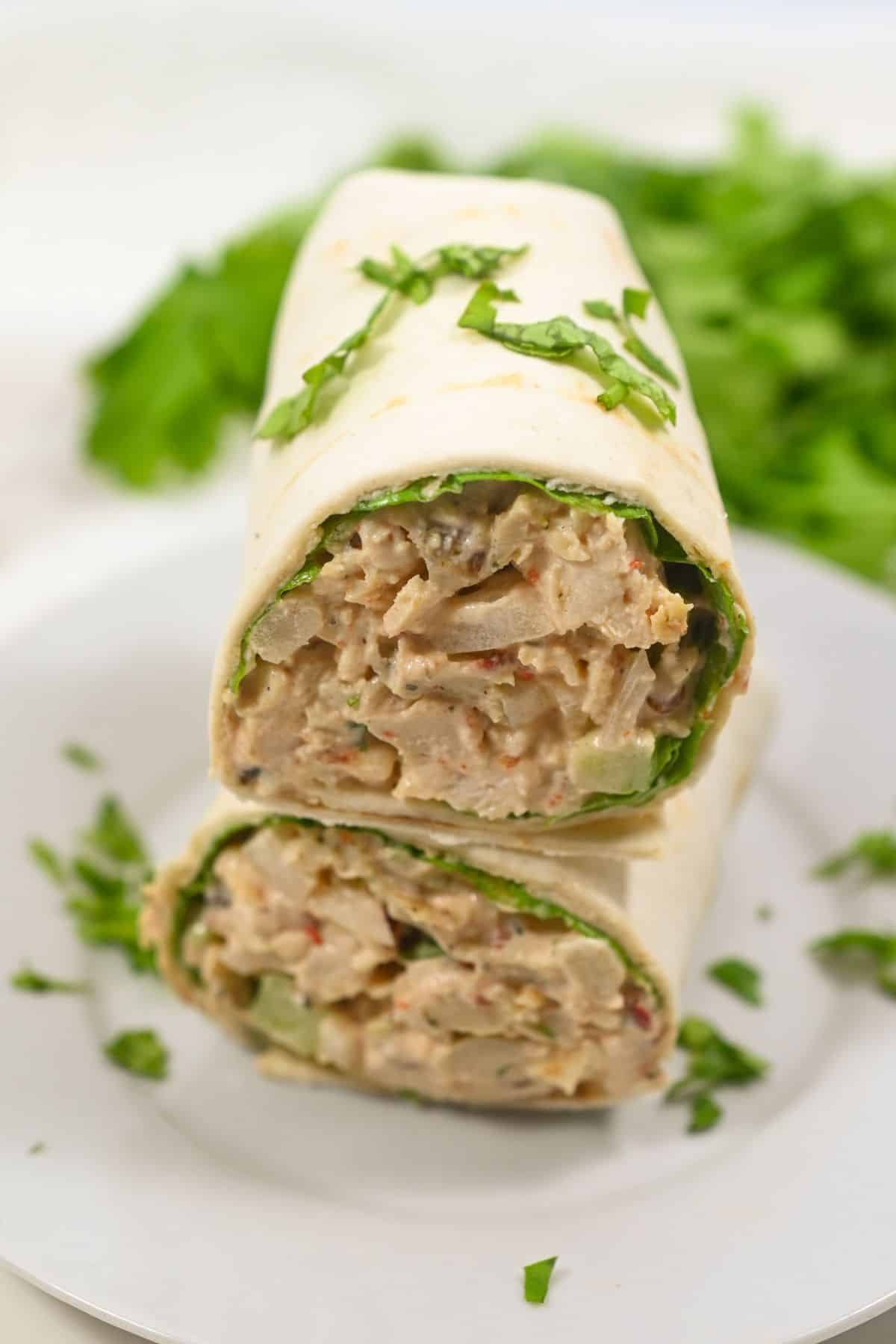 Two halves of a chipotle chicken salad wrap stacked on a white plate, garnished with chopped parsley, with leafy greens in the background.