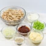 Seven glass bowls containing the flavorful ingredients mayonnaise, sour cream, chopped celery, chopped onion, chipotle peppers in sauce, and seasoning—are artfully arranged on a white surface.