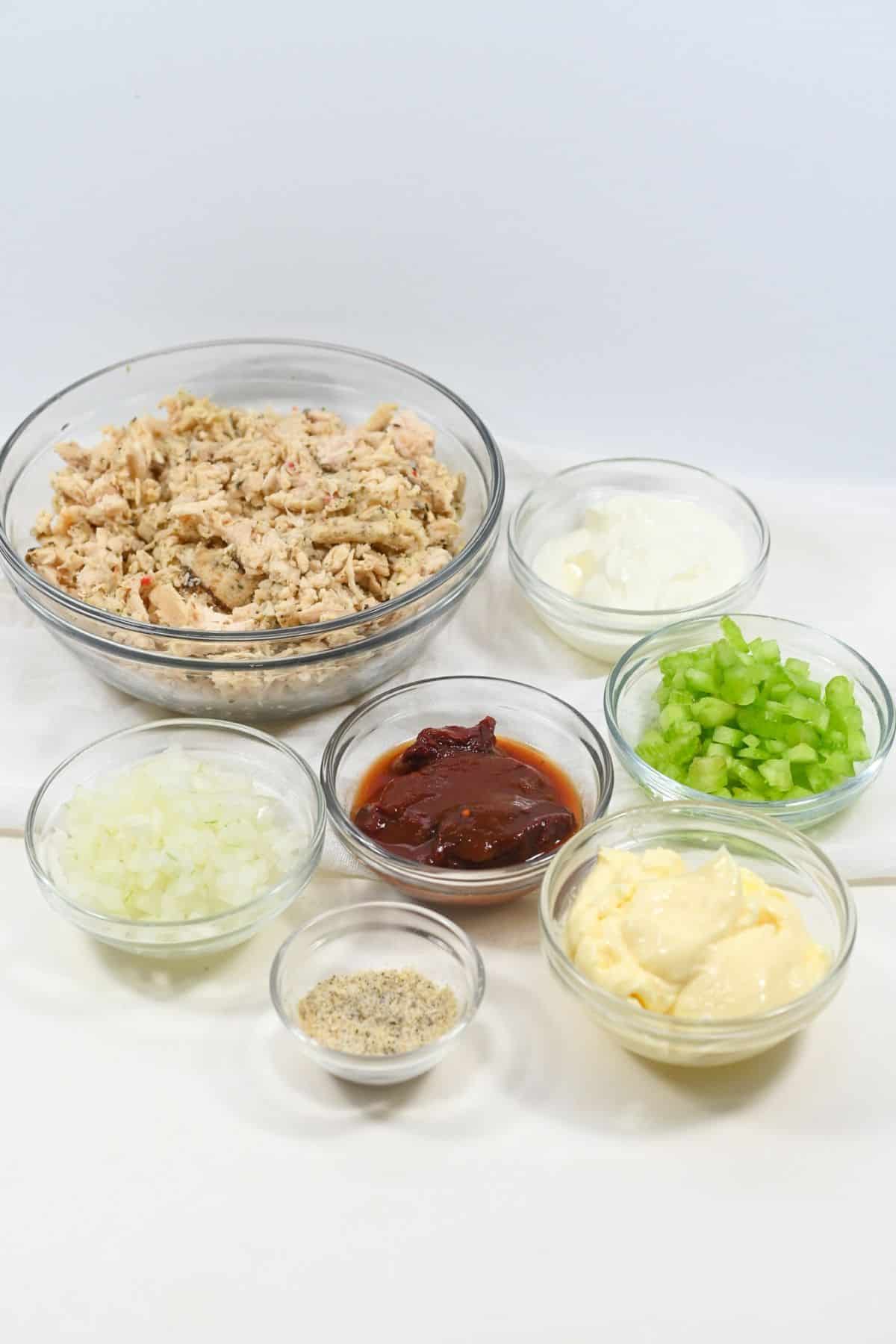 Seven glass bowls containing the flavorful ingredients mayonnaise, sour cream, chopped celery, chopped onion, chipotle peppers in sauce, and seasoning—are artfully arranged on a white surface.