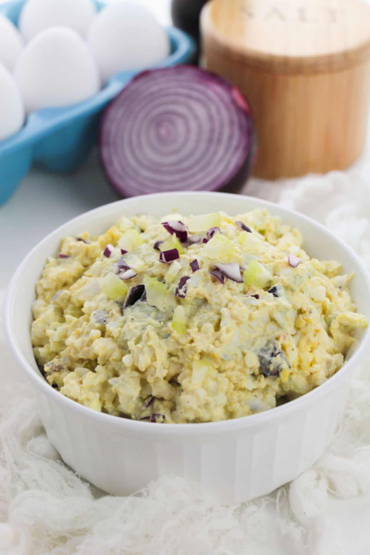 A bowl of Classic Egg Salad topped with chopped red onion, with a sliced red onion, a carton of eggs, and a salt container in the background.