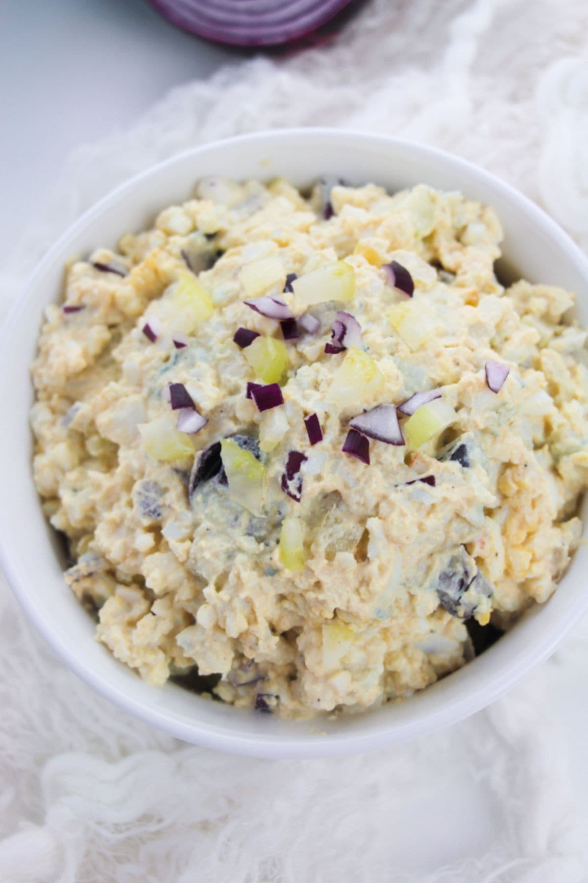 A bowl of Classic Egg Salad topped with chopped red onions, served on a white surface.