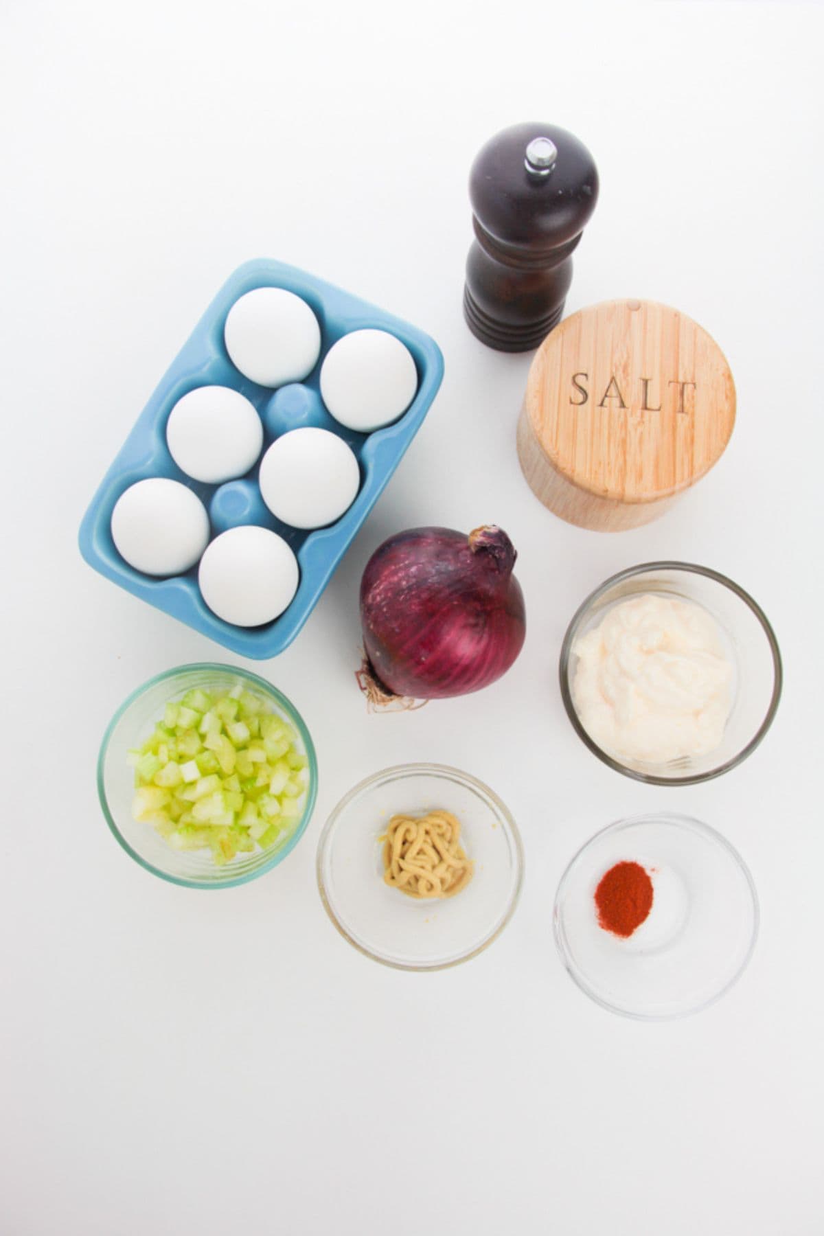 Ingredients on a white surface include eggs, black pepper, salt, red onion, celery, mayonnaise, Dijon mustard, paprika, and granulated sugar.