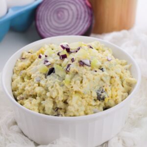 A white bowl filled with Classic Egg Salad topped with chopped red onion, with a sliced red onion and eggs visible in the background.