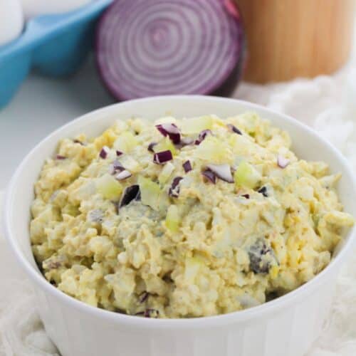 A white bowl filled with Classic Egg Salad, topped with chopped red onion, sits in front of a red onion, a salt container, and a carton of eggs.