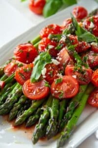 A plate of Cold Asparagus Salad topped with halved cherry tomatoes, fresh basil leaves, chopped chives, and crumbled cheese.