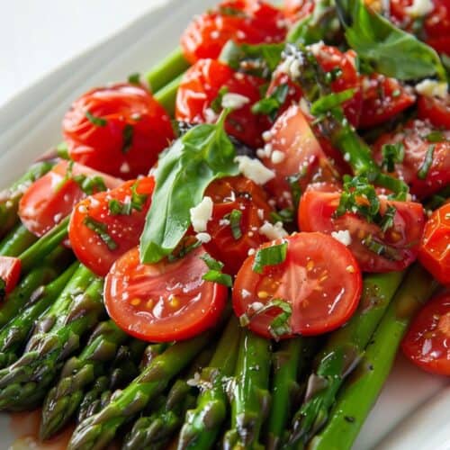 A plate of Cold Asparagus Salad topped with halved cherry tomatoes, fresh basil leaves, chopped chives, and crumbled cheese.