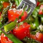 A close-up of a fork holding Cold Asparagus Salad with asparagus spears, diced tomatoes, chopped herbs, and crumbled cheese.