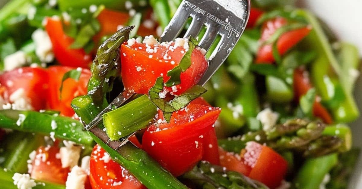 A close-up of a fork holding Cold Asparagus Salad with asparagus spears, diced tomatoes, chopped herbs, and crumbled cheese.