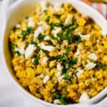 A white dish filled with Elote Salad—Mexican street corn topped with crumbled cheese and chopped herbs—rests on a marble surface.