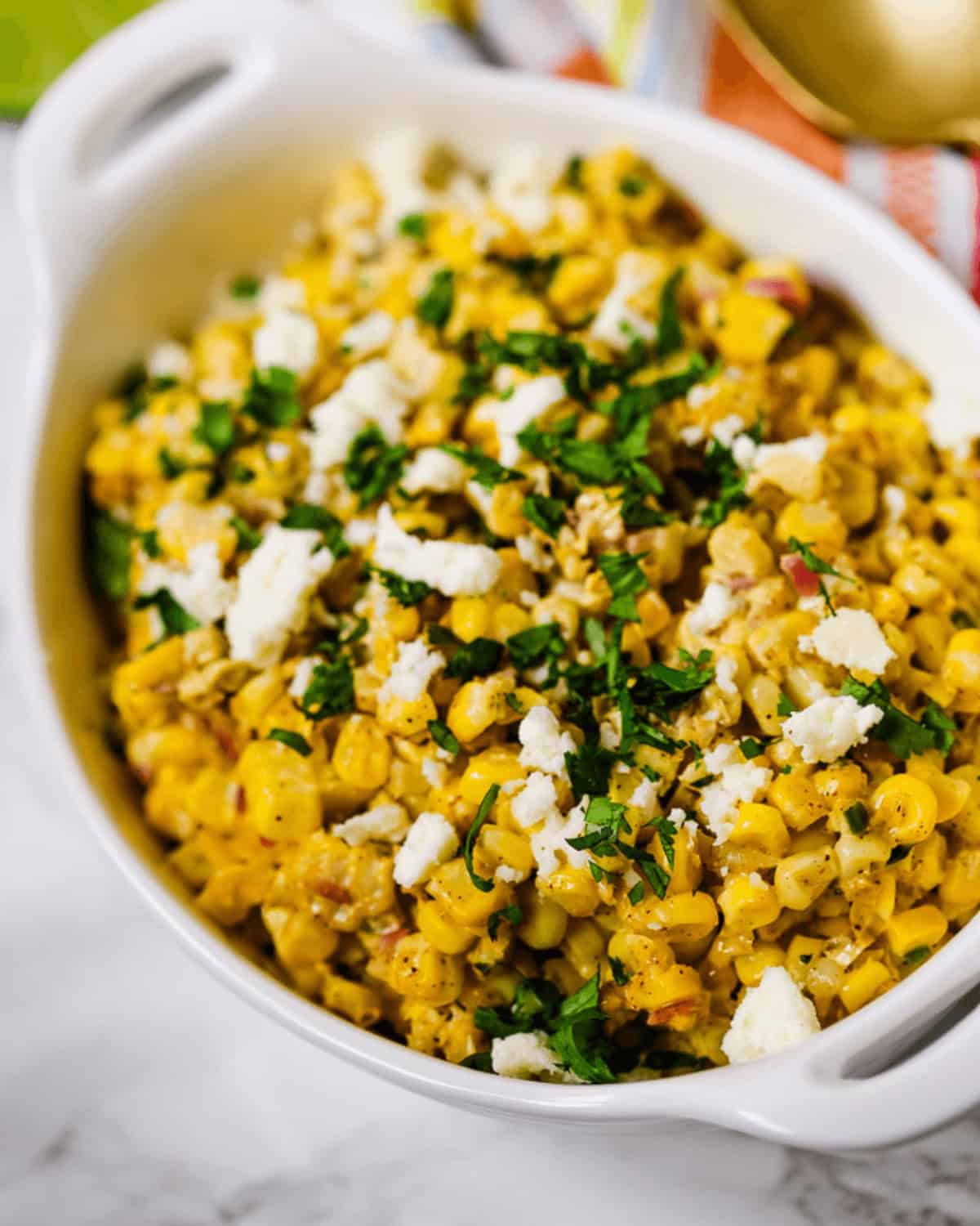 A white dish filled with Elote Salad—Mexican street corn topped with crumbled cheese and chopped herbs—rests on a marble surface.