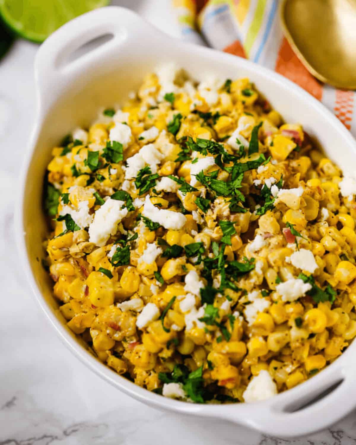 A white casserole dish filled with seasoned Elote Salad—corn topped with crumbled cheese and chopped herbs—sits on a marble surface.