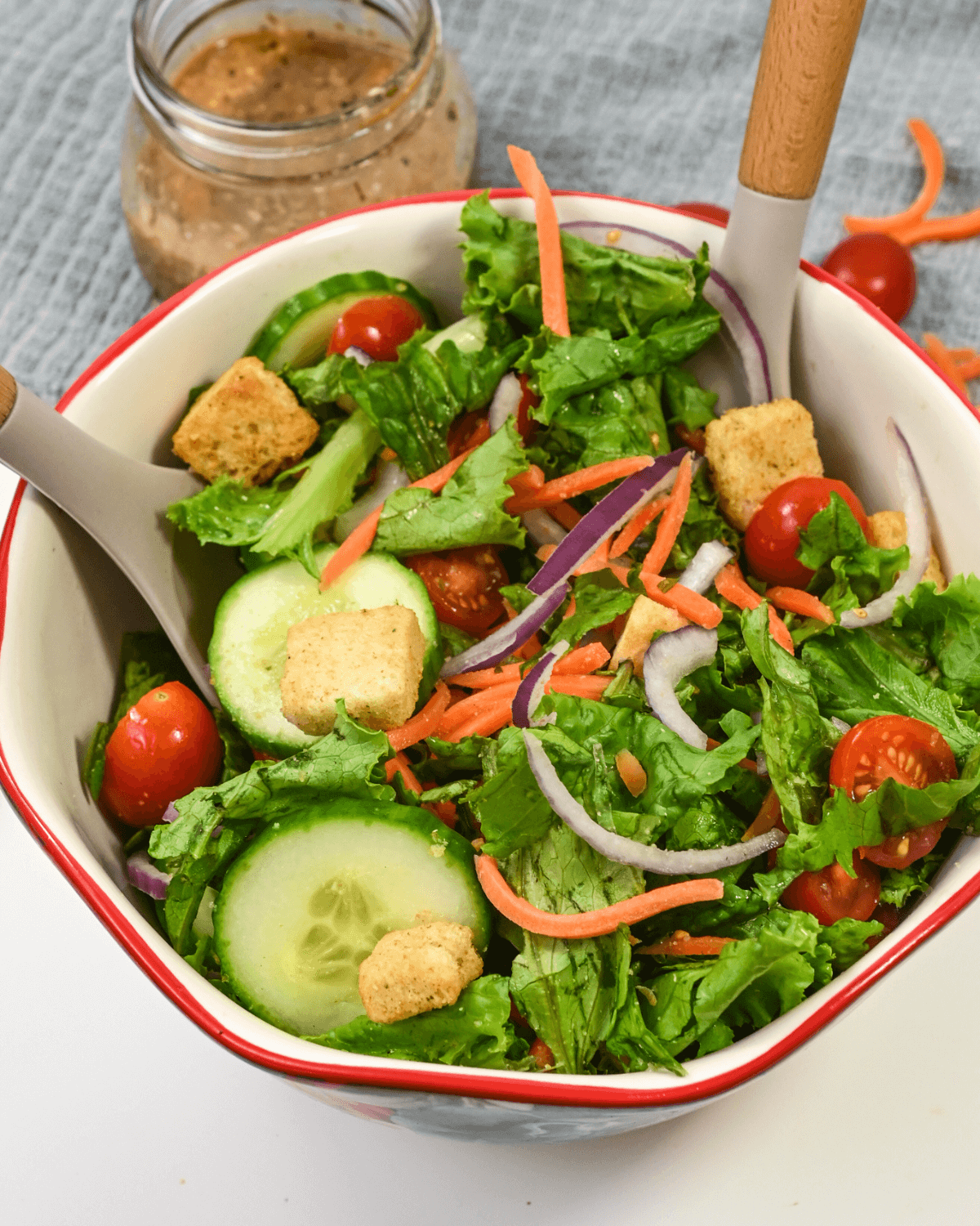 A bowl of fresh house salad with lettuce, cherry tomatoes, shredded carrots, cucumbers, red onion, and croutons, served with a side of creamy balsamic dressing.