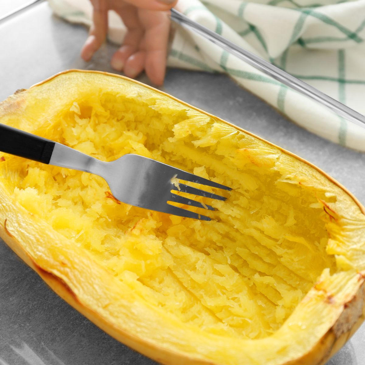 A fork fluffs the inside of a cooked spaghetti squash on a kitchen counter for a recipe index, with a person's hand visible.