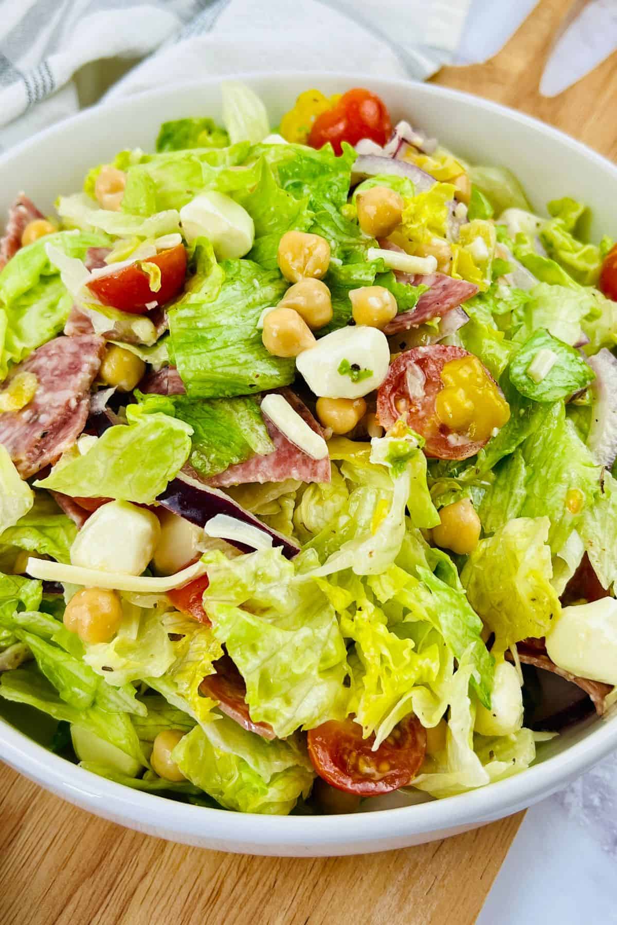 A bowl of Italian Chopped Salad with lettuce, chickpeas, cherry tomatoes, mozzarella balls, salami slices, and yellow peppers sits on a wooden board.