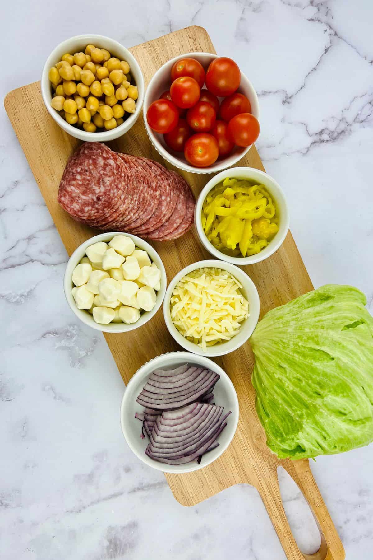 A wooden board with bowls of chickpeas, cherry tomatoes, sliced salami, mozzarella balls, shredded cheese, banana peppers, red onion, and a wedge of iceberg lettuce—everything you need to create a vibrant Italian Chopped Salad.