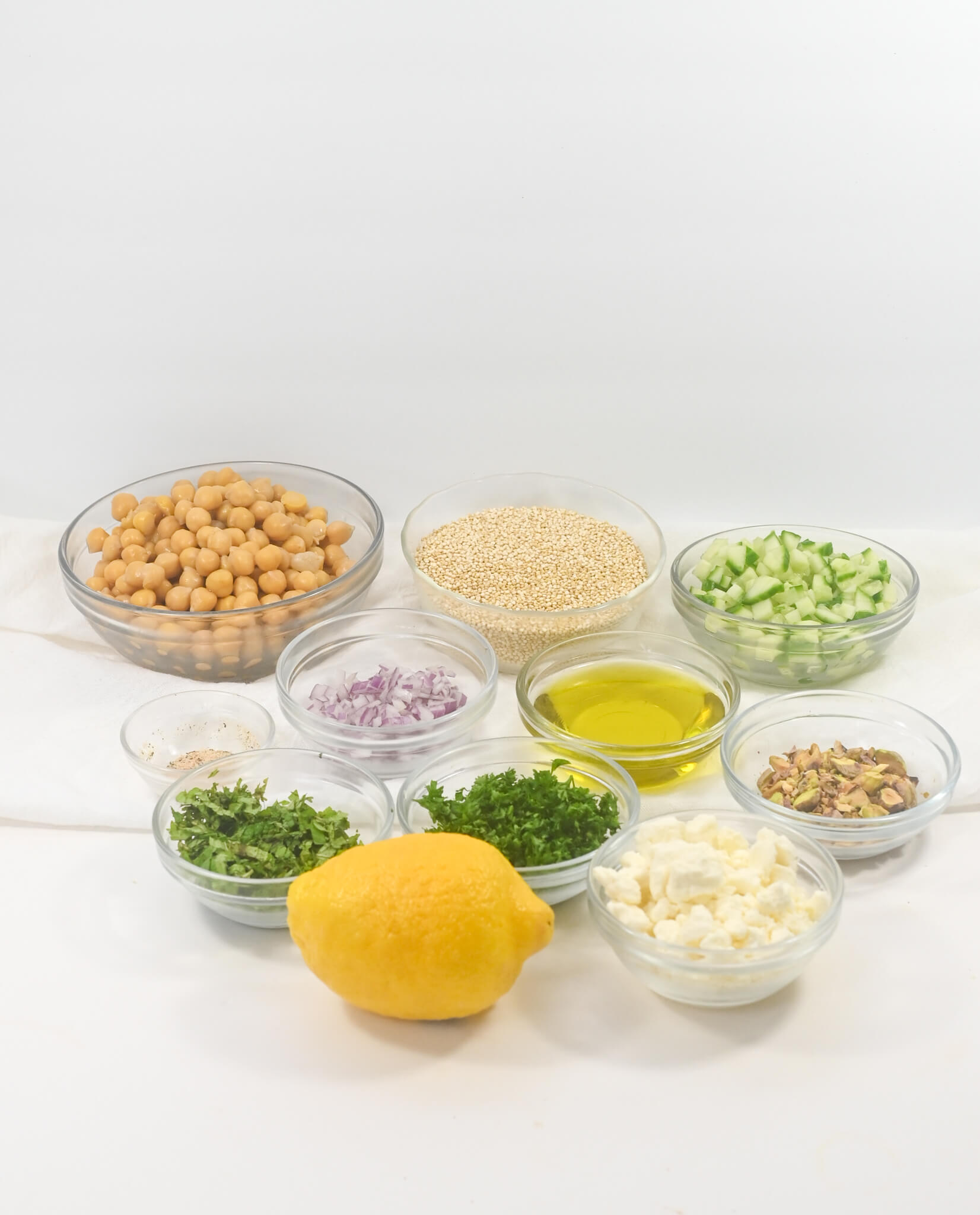 Ingredients for the dish salad arranged neatly, including chickpeas, quinoa, cucumbers, red onions, herbs, lemon, nuts, oil, and feta cheese in separate