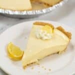 A slice of creamy Lemon Icebox Pie with a graham cracker crust, topped with whipped cream and a lemon wedge, sits on a white plate; the rest of the pie is visible in the background.