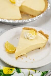 A slice of creamy Lemon Icebox Pie with a graham cracker crust, topped with whipped cream and a lemon wedge, sits on a white plate; the rest of the pie is visible in the background.