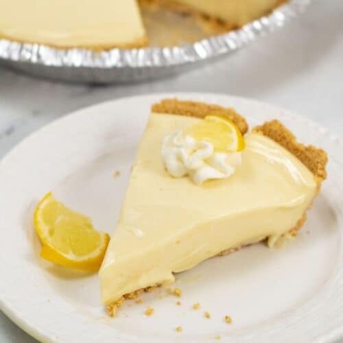 A slice of creamy Lemon Icebox Pie with a graham cracker crust, topped with whipped cream and a lemon wedge, sits on a white plate; the rest of the pie is visible in the background.